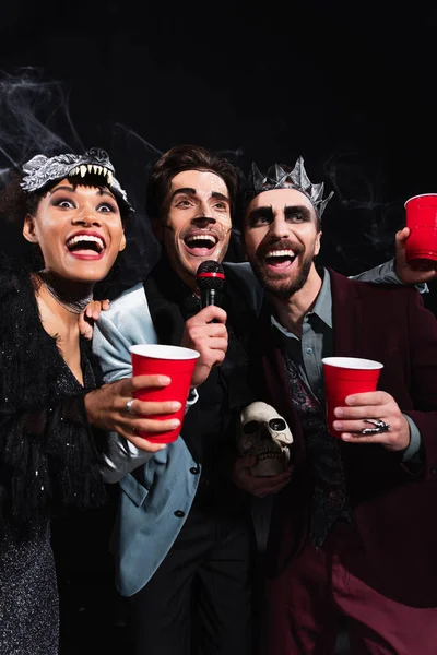 Happy multiethnic friends in halloween costumes singing karaoke on black — Stock Photo