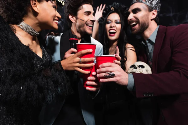 Alegres amigos multiétnicos brindando con copas de plástico mientras cantan karaoke en negro - foto de stock