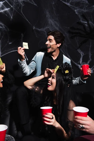 Cheerful man looking at sticky note while playing guess who game with multiethnic friends on halloween party on black — Stock Photo