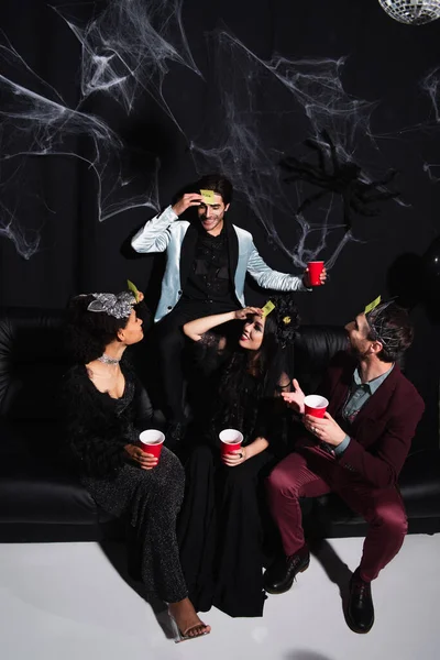 Amigos multiétnicos alegres em trajes de Halloween jogando adivinhem quem jogo no sofá no preto — Fotografia de Stock