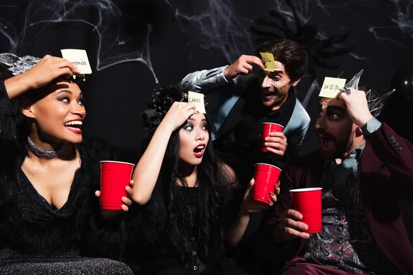 Astonished asian woman near cheerful multiethnic friends playing guess who game on halloween party on black — Stock Photo