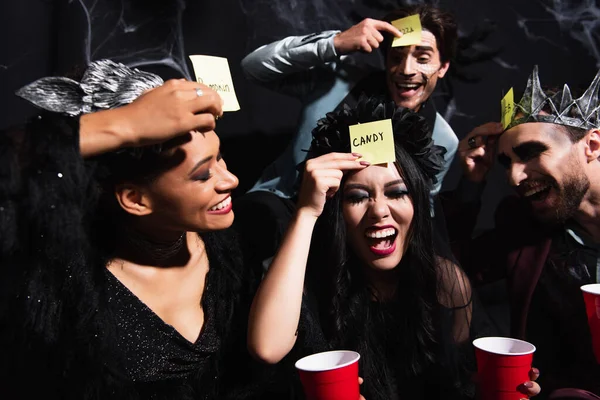 Excited interracial women laughing with closed eyes while playing guess who game on halloween party on black — Stock Photo