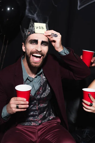 Homem em vampiro rei coroa jogando adivinhem quem jogo durante festa de Halloween com amigos em preto — Fotografia de Stock