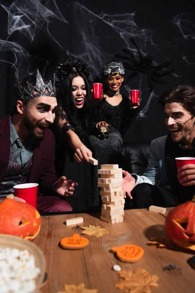 Mujer afroamericana emocionada jugando juego de torre de madera con amigos multiétnicos en trajes de Halloween en negro - foto de stock