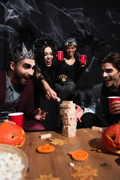 Animado Africano americano mulher jogando jogo de torre de madeira com amigos multiétnicos em trajes halloween em preto — Fotografia de Stock