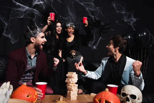 Man showing win gesture near wood blocks game while interracial women toasting with plastic cups on black — Stock Photo