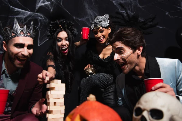 Emocionado mujer asiática en traje de Halloween vampiro jugando bloques de madera juego con amigos multiétnicos en negro - foto de stock