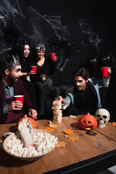 Man in halloween makeup playing wood blocks game with multiethnic friends near carved pumpkins and popcorn on black — Stock Photo
