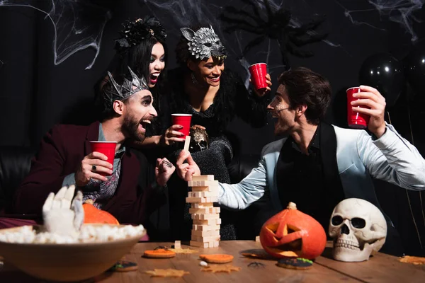 Hombre alegre con el maquillaje de Halloween jugando bloques de madera juego con amigos multiétnicos en negro - foto de stock