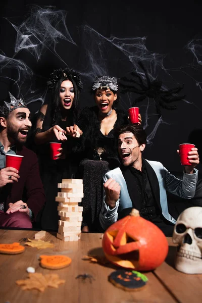 Excited man in halloween makeup showing win gesture near wood blocks game and multiethnic friends on black — Stock Photo