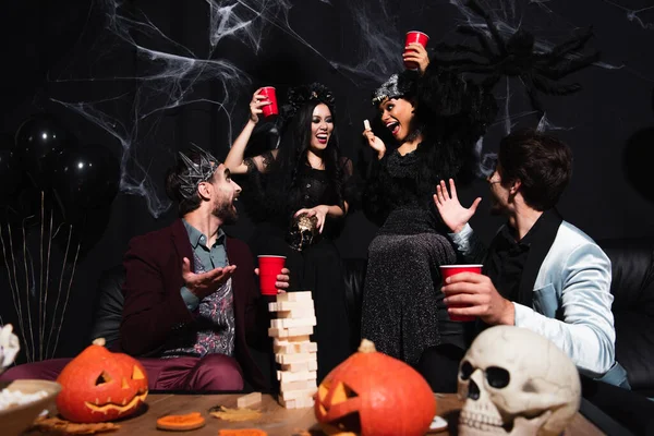 Men pointing with hands near wood blocks game while cheerful interracial women in halloween costumes toasting with plastic cups on black — Stock Photo