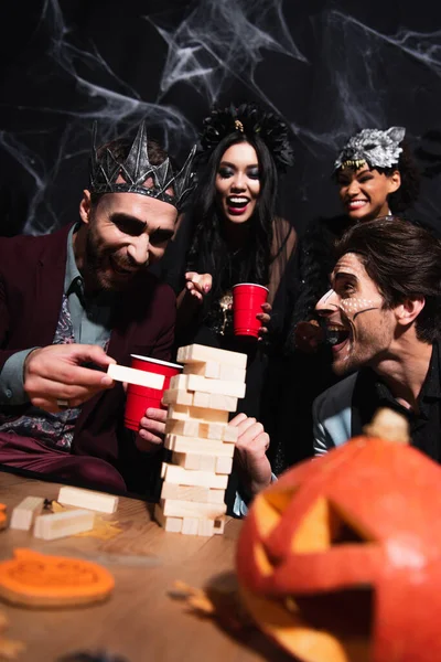 Cheerful man in vampire king crown playing wood blocks game during halloween party with multiethnic friends on black — Stock Photo