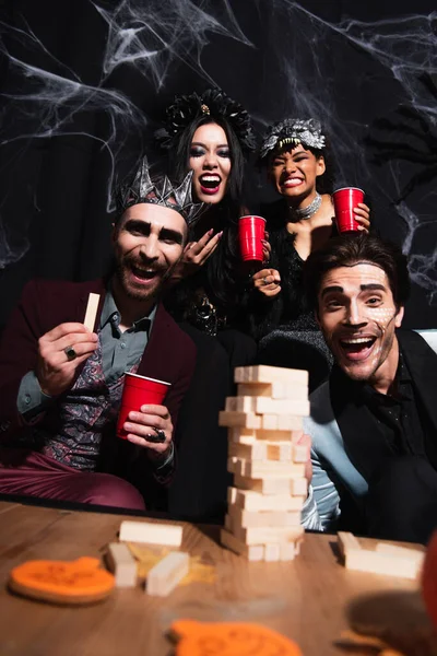 Excited multiethnic friends in halloween costumes looking at camera near wood blocks game on black — Stock Photo