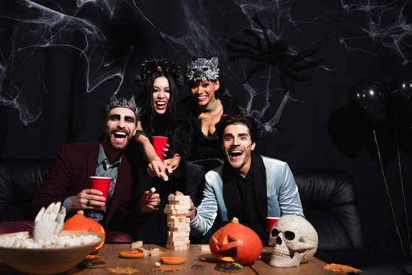 Excited multicultural women in halloween costumes playing wood blocks game with men on black — Stock Photo
