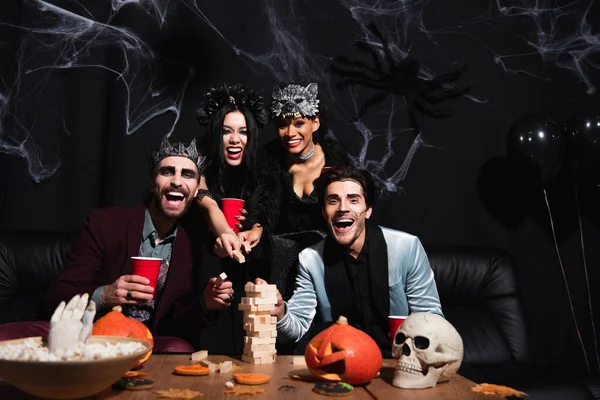 Animado multicultural mulheres em halloween trajes jogar madeira blocos jogo com homens no preto — Fotografia de Stock