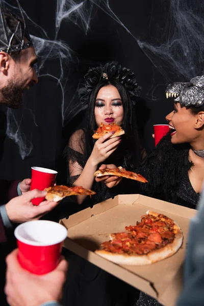 Pleased asian woman in vampire halloween costume eating pizza near multiethnic friends on black — Stock Photo