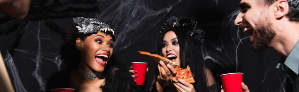 Thrilled african ameican woman holding pizza during halloween party with multiethnic friends on black, banner — Stock Photo