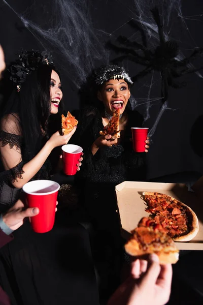 Excited interracial women in halloween costumes eating pizza during party on black — Stock Photo