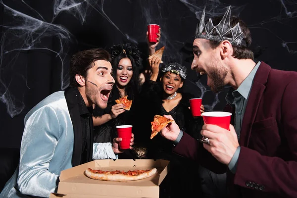 Erstaunte Männer, die ihren Freund in der Nähe einer Pizza anschauen und aufgeregte interrassische Frauen in Halloween-Kostümen auf schwarz — Stockfoto