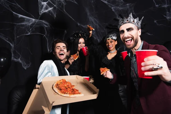 Збуджений чоловік у макіяжі на Хелловін показує піцу біля щасливих багатоетнічних друзів на чорному — стокове фото