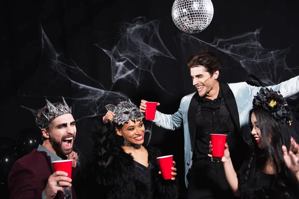 Cheerful african american woman adjusting wolf mask while dancing on halloween party with interracial friends on black — Stock Photo