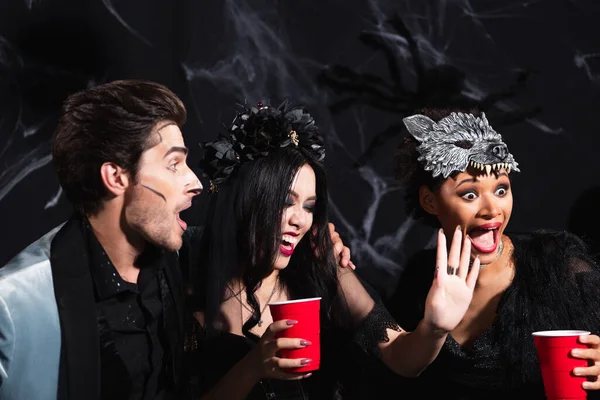 Sorrindo asiático mulher mostrando parar gesto perto emocionante multiétnico amigos durante halloween festa no preto — Fotografia de Stock