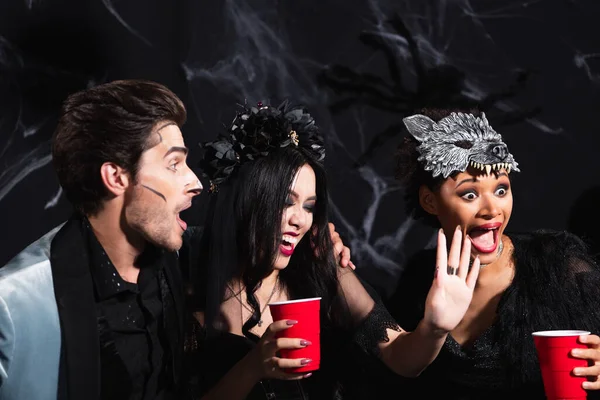 Sonriente asiático mujer mostrando parada gesto cerca emocionado multiétnico amigos durante halloween fiesta en negro - foto de stock