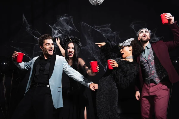 African american woman in halloween costume pointing with finger while dancing with excited multiethnic friends on black — Stock Photo