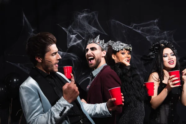 Fröhliche multiethnische Freunde mit Plastikbechern, die auf einer Halloween-Party auf Schwarz tanzen — Stockfoto