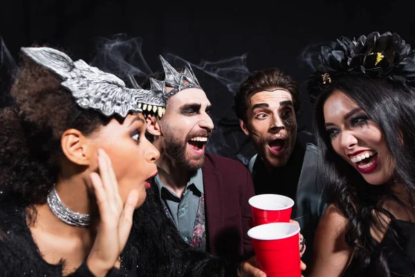 Excitée femme afro-américaine en masque de loup parler à des amis multiethniques étonnés pendant la fête sur noir — Photo de stock