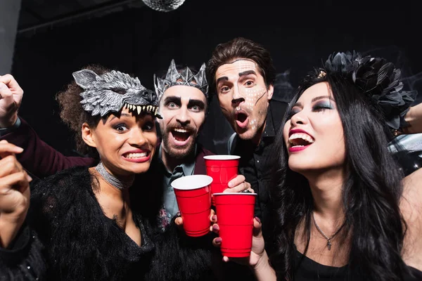 Excited multiethnic friends looking at camera while toasting with plastic cups on black — Stock Photo