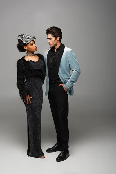 Чоловік з рукою в кишені дивиться на афроамериканську жінку в чорному платті і перевертень маску на сірому — стокове фото