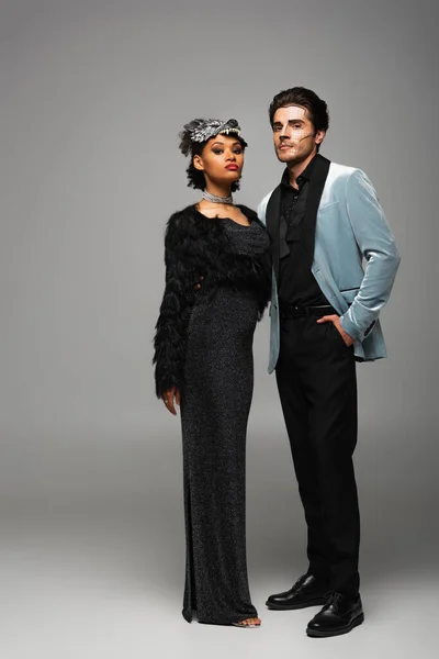 Seguro y elegante pareja interracial en disfraces de Halloween mirando a la cámara en gris - foto de stock