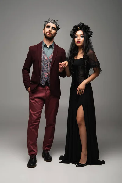 Elegante interracial pareja en vampiros halloween trajes cogidos de la mano mientras de pie en gris - foto de stock