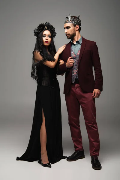 Élégante femme asiatique en robe noire et couronne embrassant épaule de l'homme en costume de roi vampire halloween sur gris — Photo de stock