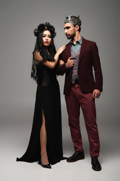 Elegante asiático mulher no preto vestido e grinalda abraçando ombro de homem no vampiro rei halloween traje no cinza — Fotografia de Stock