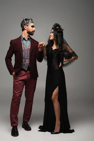 Man in vampire king halloween costume holding hand of asian woman in black dress on grey — Stock Photo