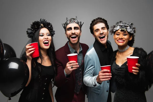 Alegres amigos multiétnicos sosteniendo copas de plástico y riendo durante la fiesta de Halloween aislados en gris - foto de stock