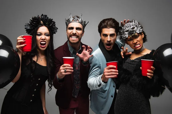 Spooky multiethnic friends with plastic cups growling and showing scary gesture isolated on grey — Stock Photo