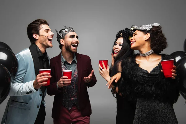 Amigos interracial emocionados sosteniendo copas de plástico y riendo durante la fiesta de Halloween aislado en gris - foto de stock