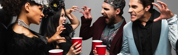 Amigos multiétnicos con copas de plástico asustándose entre sí durante la fiesta de Halloween aislados en gris, pancarta - foto de stock