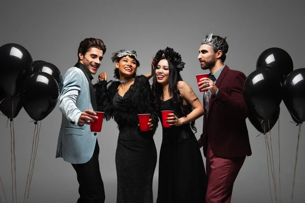 Happy multiethnic friends in halloween costumes holding plastic cups near black balloons isolated on grey — Stock Photo