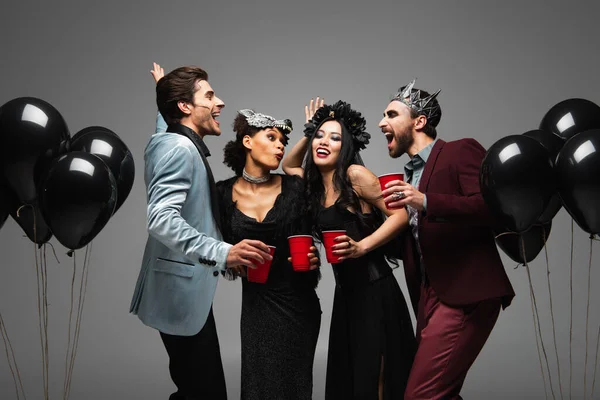 Багатоетнічні друзі з пластиковими чашками позують біля чорних кульок на вечірці Хеллоуїна ізольовані на сірому — стокове фото