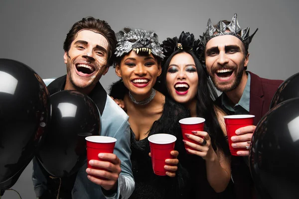 Cheerful multiethnic friends in halloween costumes looking at camera while holding plastic cups isolated on grey — Stock Photo