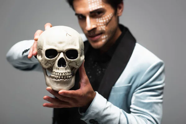Расплывчатый человек в макияже Хэллоуина показывает страшный череп, глядя на камеру, изолированную на сером — стоковое фото