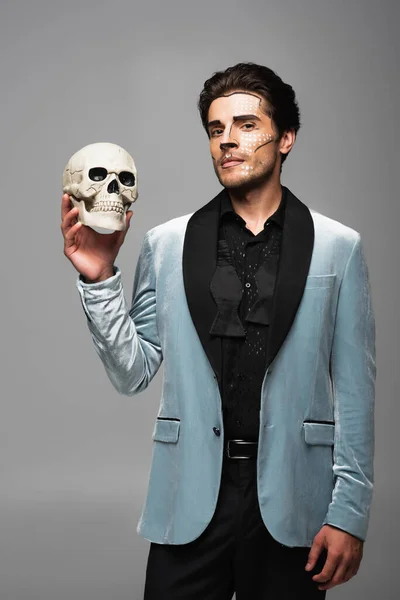 Elegant man in elegant blazer and halloween makeup holding spooky skull isolated on grey — Stock Photo