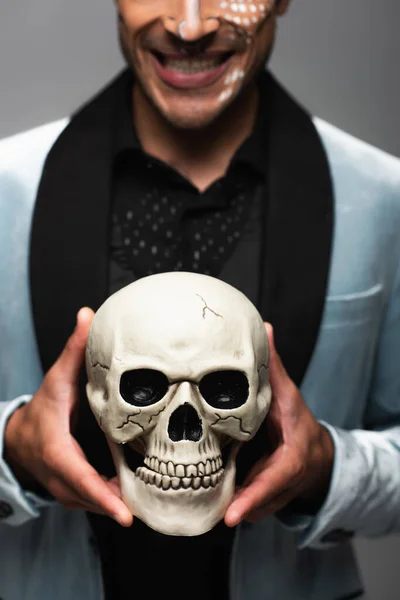 Abgeschnittene Ansicht eines verschwommenen Mannes mit sarkastischem Lächeln, der gruseligen Schädel isoliert auf grau hält — Stockfoto