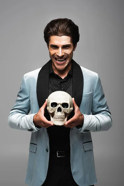 Hombre sarcástico en halloween maquillaje celebración espeluznante cráneo y riendo aislado en gris - foto de stock