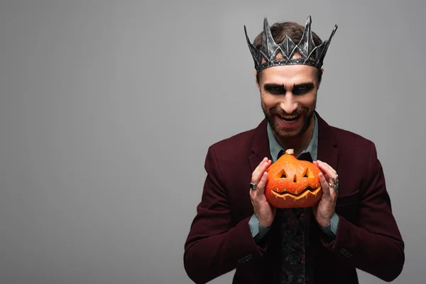 Hombre sarcástico en rey vampiro halloween corona sonriendo y mostrando espeluznante calabaza aislado en gris - foto de stock