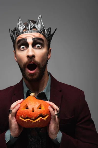 Sorprendido hombre en vampiro rey halloween corona celebración tallada calabaza aislado en gris - foto de stock